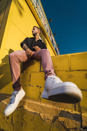 A low angle shot of a hot man wearing a black oversize shirt and pink pants sitting on yellow wallの写真素材