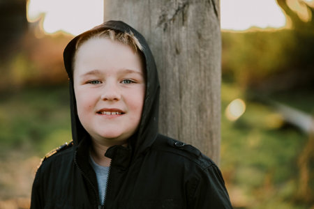 A cute blonde child wearing a black jacket and a hood and posing while leaning on a wooden pillar