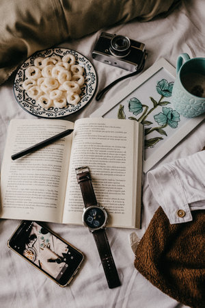 A vertical top view of an open book with a camera, coffee, and snacks on a bed
