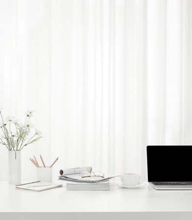 A vertical shot of a cup of coffee, a laptop,  a vase full of flowers, magazines on a white table