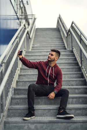 A young male wearing a red jacket with dark jeans and taking a selfie with a phone while sitting on the stairs outdoorsの写真素材