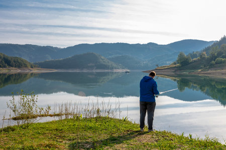 A caucasian man fishing from a beautiful calm lake, surrounded by green hills during daylightの写真素材