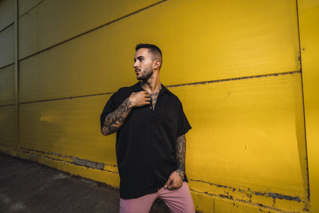 A young hot man wearing a black oversize shirt and pink pants standing against a bright yellow wall