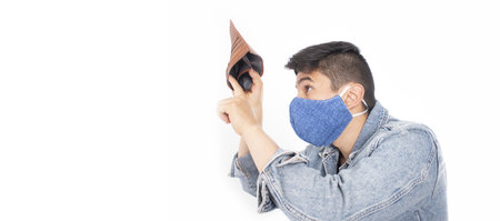 A closeup of a young man wearing a facemask, looking at his empty wallet in front of a white...