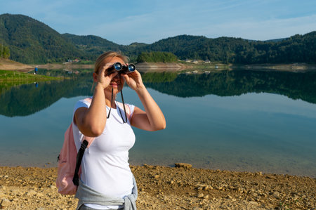 A caucasian woman enjoying the view with binoculars, standing near a beautiful calm lake, surrounded by green hillsの写真素材