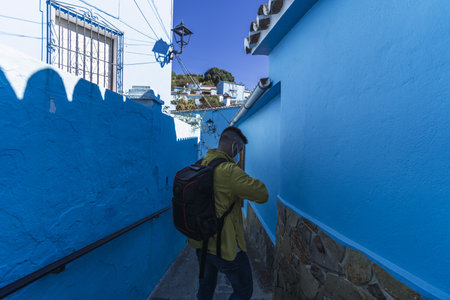 A young male with a backpack wearing a yellow jacket and walking through an alley with blue wallsの写真素材