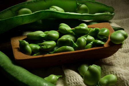A closeup of fresh raw broad beans in the pods and a pile of beans in the wooden dishの写真素材