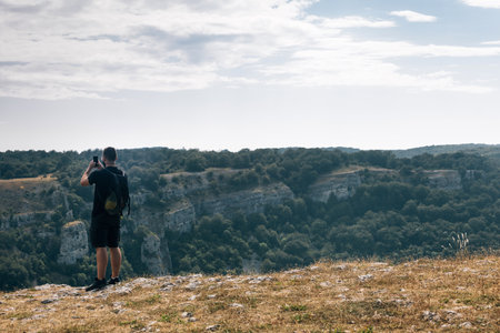 A male hiker taking a picture of hills covered in greenery with his phone under a cloudy skyの写真素材