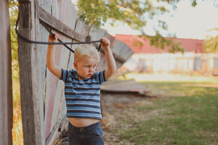 A blonde little boy playing with a rope hanging from an old wooden fenceの写真素材