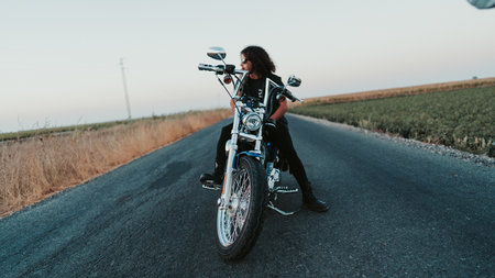A shallow focus of a cool curly-haired male posing on his motorcycle on the roadの写真素材