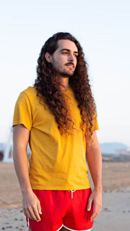 A vertical shot of a young man with long curly hair standing on the beachの写真素材