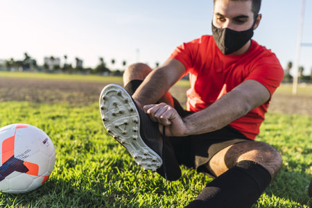 A male footballer in a mask sitting on the lawn and putting on a sneaker, the concept of a healthy lifestyle in "new normal" pandemic conditionsの写真素材