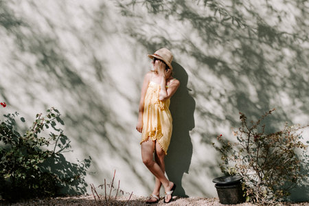 A beautiful young lady wearing a beach hat, beach dress, and sunglasses posing leaning on a wallの写真素材