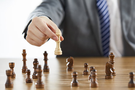 A businessman in a suit using his white king chess piece among dark chess pieces on a table -...