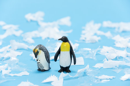 A closeup shot of penguin toys surrounded with pieces of plastic and paper on a blue backgroundの写真素材