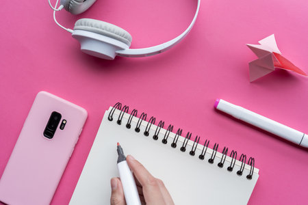 A high angle shot of a cellphone, headphones, and writing tools on a pink surfaceの写真素材