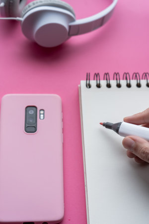 A vertic shot of a cellphone, headphones, and writing tools on a pink surface