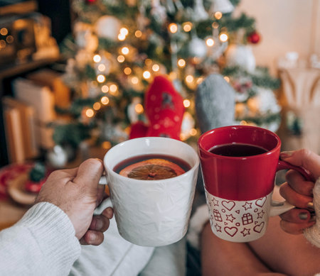 A selective focus shot of a couple in knitted socks drinking hot tea with a glowing bright Christmas tree in the backgroundの写真素材