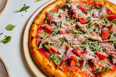 A closeup shot of a delicious cheesy pizza with ham, tomatoes, and greens on a plateの写真素材