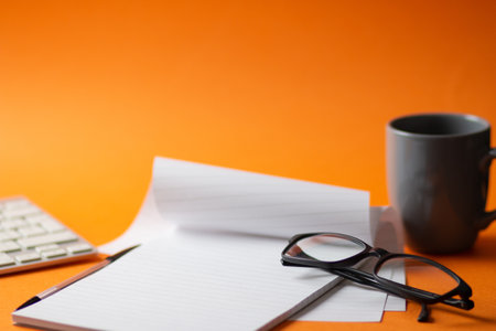 A selective focus shot of a keyboard, notepad, pen and a cup of hot coffee isolated on an orange background - work from home conceptの写真素材