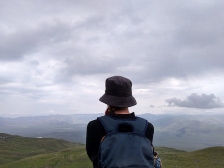 A shallow focus of a hiker with a backpack and a bucket hat standing on a hill under the cloudy sky