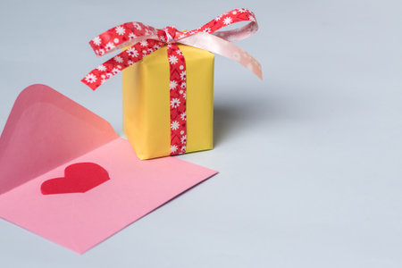Minimal composition with a pink envelope with red heart inside, and a yellow gift box with a red...