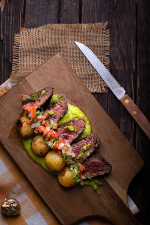 A vertical shot of beef steaks with roasted potatoes, sauce, and vegetable salad on a wooden boardの写真素材
