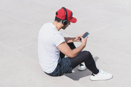 A young sportsman in headphones  sitting on the street and looking at his phoneの写真素材