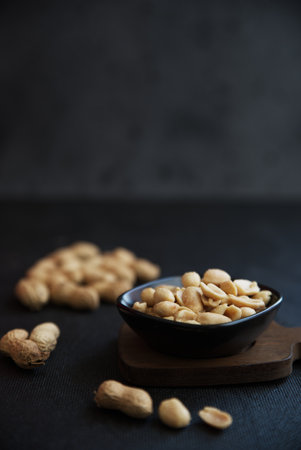 A closeup shot of a tiny black bowl on a wooden board filled with peanuts on a black background