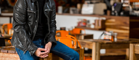 A selective focus shot of a male wearing a black shirt, leather jacket, and jeans sitting on a tableの写真素材