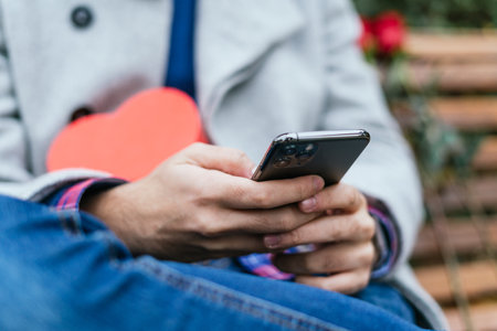 A selective focus closeup of a male holding a mobile phone with a red heart on his lap on a bench outdoors-concept Valentine's dayの写真素材