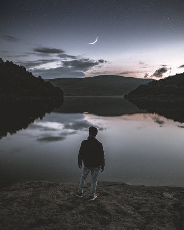 Man in black jacket and red shoes standing in front of a lake and a sunset sky with a moon during duの写真素材
