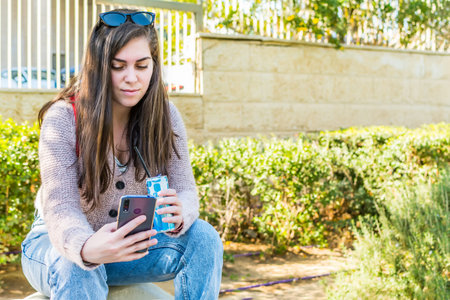 A side view of a smiling female using a smartphone with soda sitting down in a parkの写真素材
