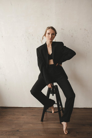 A beautiful young European girl in a black suit posing at the camera while sitting on a stoolの写真素材