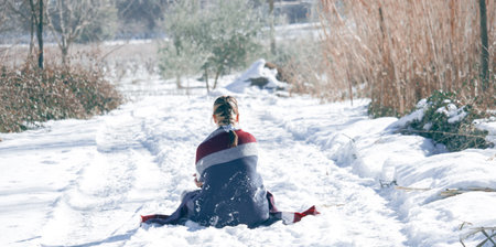 A selective focus shot of a lady covered in a winter scarf sitting in a snowy field