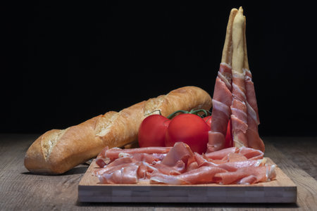 Spanish ham on a wooden plate next to some bread-sticks wrapped in ham