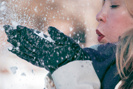 A selective focus shot of a beautiful young lady blowing the snow on her hands wearing glovesの写真素材