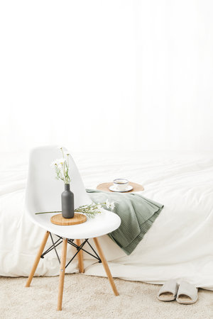 A vertical shot of a simple chair near a bed with a cup of coffee on it against a white wallの写真素材