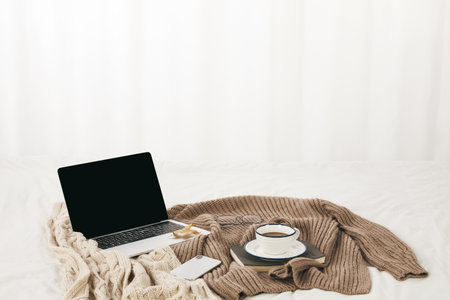 A closeup of a modern laptop on knitted sweaters with a cup of coffee on a notepad on themの写真素材