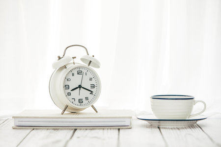 A closeup of an alarm clock on a notepad with a cup of coffee on the table against a white...