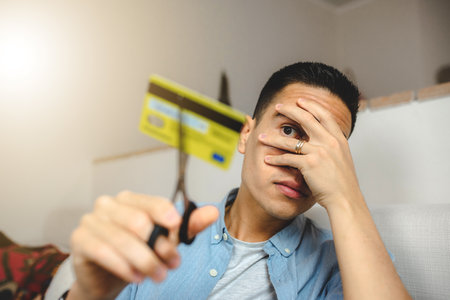 A young Hispanic male cutting credit card with scissors, scared covering face with hand - Internet banking and going bankrupt conceptの写真素材