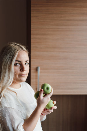 A vertical shot of a blonde woman holding apples in the kitchenの写真素材
