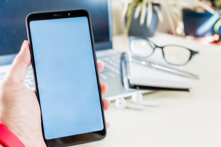 A closeup of a person holding a smartphone with a white screen above a deskの写真素材