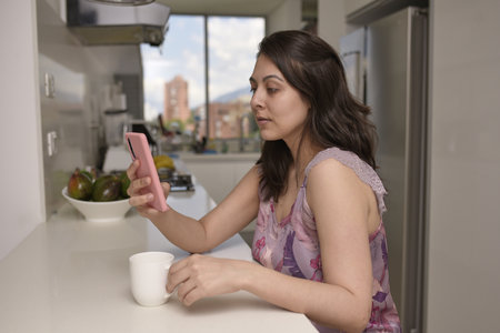 A closeup portrait of a Hispanic woman in pajamas enjoying a morning coffee in a kitchen and surfing on internetの写真素材
