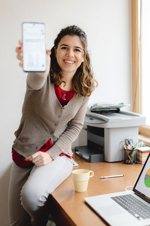 A shallow focus of a young female sitting on an office desk and showing a business report on her phoneの写真素材