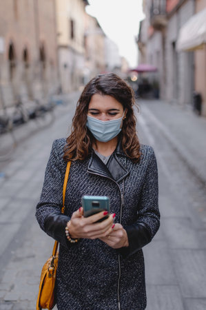 A shallow focus of a young brunette woman with a facemask using her phone outdoors - COVID-19の写真素材
