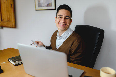 A handsome young man with a bright smiling face looking at the camera while holding a tablet with a laptop in front of himの写真素材