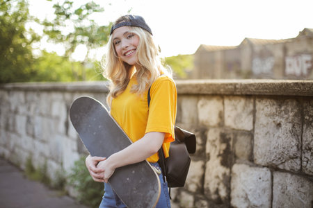 A shallow focus of a young smiling blonde woman with a skateboard posing in a parの写真素材