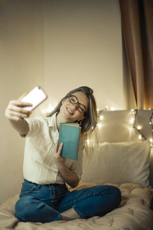 A shallow focus of a woman with glasses sitting on her bed and taking a selfie with a bookの写真素材