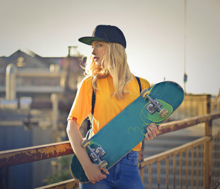 A shallow focus of a blonde woman wearing a yellow t-shirt with jeans and a snapback and posing with a skateboardの写真素材